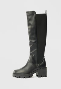 Botas negras hasta la rodilla con una combinación de cuero suave y tela texturizada, que presentan una suela gruesa y un tacón cuadrado para mayor estabilidad y agarre.