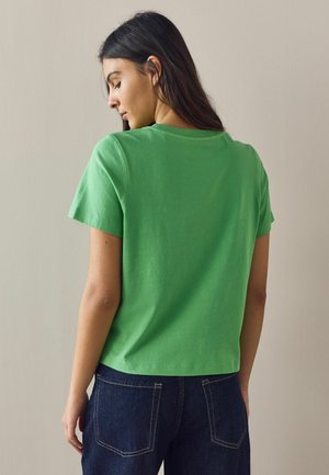 Frau mit langen dunklen Haaren, die ein schlichtes grünes Kurzarmshirt und dunkelblaue Jeans trägt, mit dem Rücken zu beigefarbenem Hintergrund.