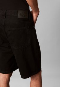 Mann trägt schwarze Calvin Klein Shorts mit Gesäßtaschen, steht vor einem schlichten hellgrauen Hintergrund.