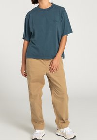 Przesadnie duży niebieski bawełniany t-shirt z krótkim rękawem, ozdobiony małym czarnym logo, w połączeniu z jasnobeżowymi bawełnianymi spodniami i białymi sneakersami.