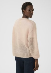 Lyserød, fluffy strikket sweater med ribbet kant og brede ærmer, der viser en blød tekstur. Båret over mørke jeans med et enkelt design.