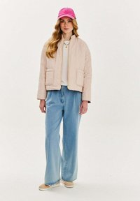 Pletená béžová bomber bunda s prešívanou textúrou, zlatý zips, dve predné vrecká; kombinovaná so svetlomodrými širokými džínami a ružovou čiapkou.