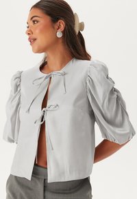 Blusa cropped grigia chiara con maniche a sbuffo, chiusura anteriore con fiocco e texture morbida. Presenta un colletto rotondo e una vestibilità leggermente ampia.