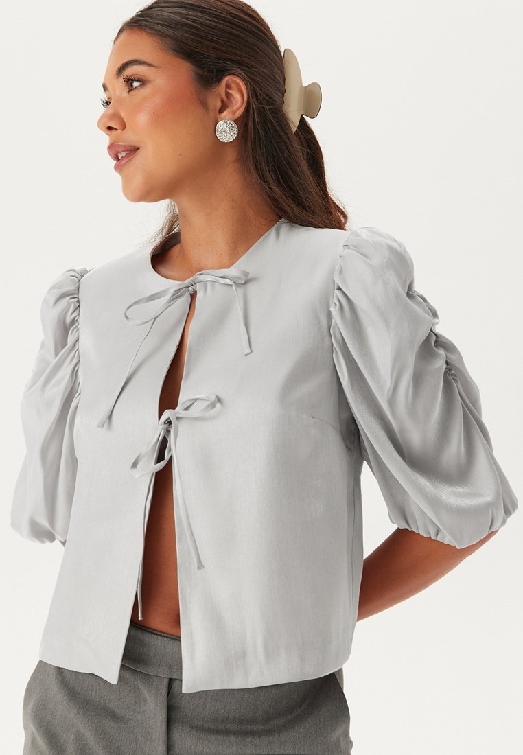 Blusa cropped grigia chiara con maniche a sbuffo, chiusura anteriore con fiocco e texture morbida. Presenta un colletto rotondo e una vestibilità leggermente ampia.