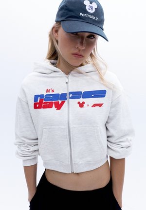 Jeune femme portant un sweat à capuche gris zippé avec le texte "It's race day" et une casquette Formula 1 bleu marine avec un logo Mickey Mouse.
