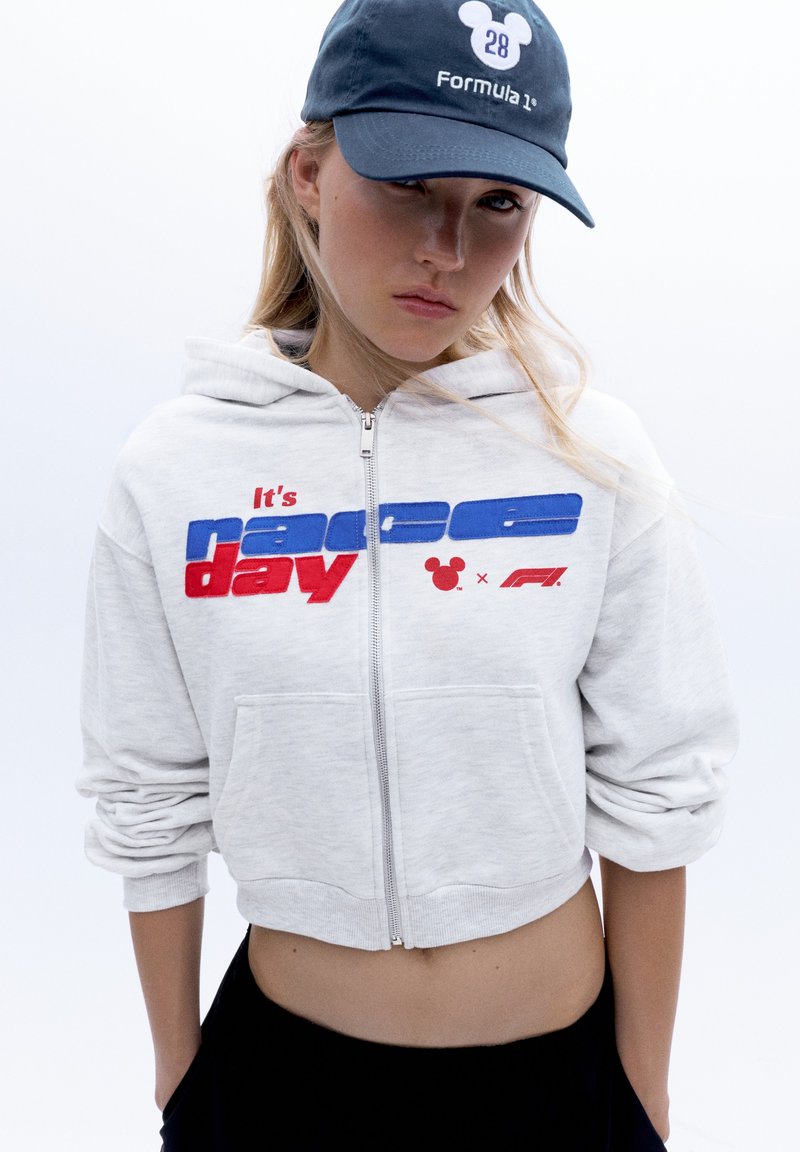 Jeune femme portant un sweat à capuche gris zippé avec le texte "It's race day" et une casquette Formula 1 bleu marine avec un logo Mickey Mouse.