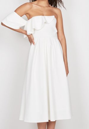Vrouw draagt een witte off-shoulder midi-jurk met een gerimpeld bovenstuk en uitlopende rok, staand met één hand op haar heup.