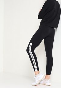 Leggings noirs en tissu extensible avec une rayure verticale blanche portant du texte sur le côté, associés à un sweat-shirt noir court et des chaussures claires.