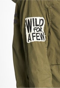 Olivgrön jacka med en framträdande vit lapp på ärmen med texten "WILD FOR A FEW" i svart, texturerad bokstav.