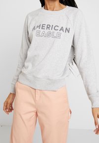Felpe grigie con la scritta "AMERICAN EAGLE" stampata in grandi lettere grasse. Presentano maniche raglan e un orlo a costine. Indossate con pantaloni pesca.