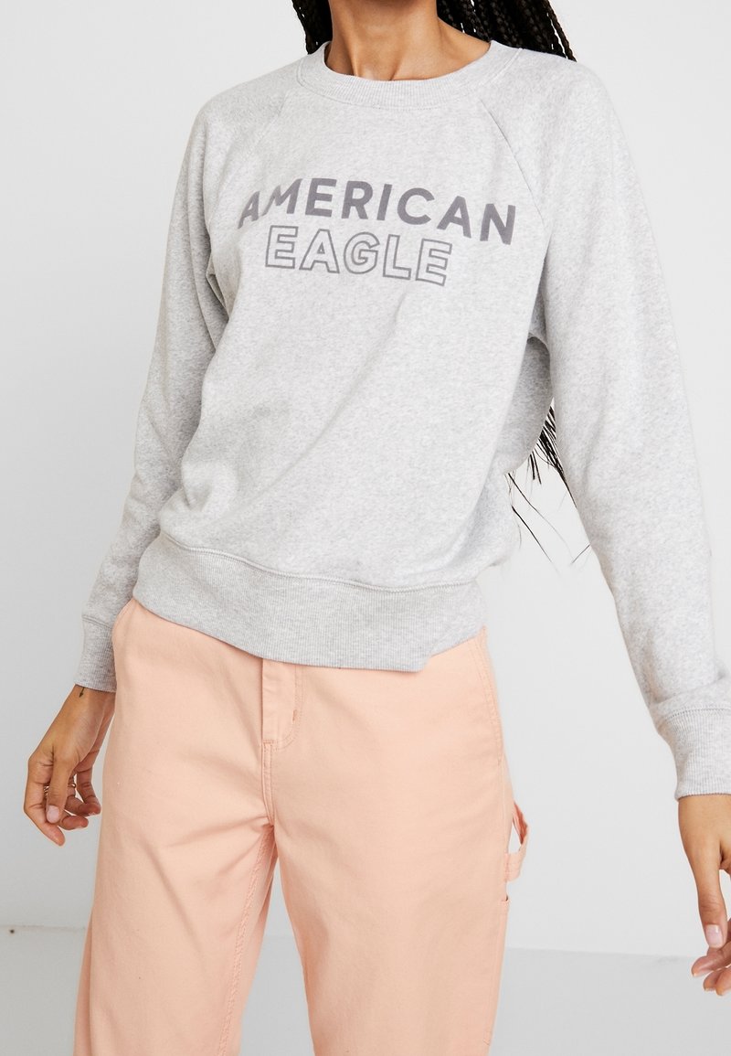 Felpe grigie con la scritta "AMERICAN EAGLE" stampata in grandi lettere grasse. Presentano maniche raglan e un orlo a costine. Indossate con pantaloni pesca.