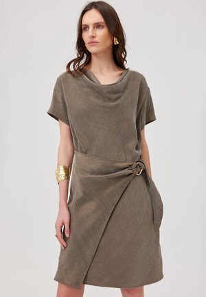 Kobieta nosząca krótką sukienkę w kolorze taupe z marszczonym dekoltem i detalem kopertowym zabezpieczonym okrągłą klamrą w talii.