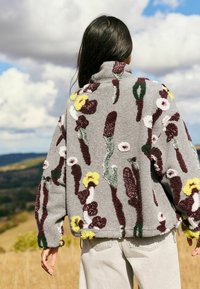 Fleecy grey jacket with floral embroidery, featuring purple, green, and yellow patterns. High collar and loose fit, with elastic hem and cuffs.