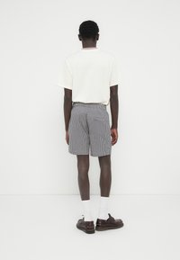 Λευκό T-shirt με κοντά μανίκια και λαιμόκοψη με ρίγες, συνδυασμένο με καρό σορτς, σκουρόκαφέ παπούτσια και λευκές κάλτσες, σε όψη από πίσω.