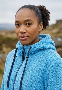 Ung kvinde med krøllet hår iført en blå fleecehættetrøje står udendørs i et naturligt, stenet landskab under en overskyet himmel.