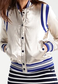Giacca bomber in satin color oro chiaro con polsini e orlo a righe blu e bianche, dotata di chiusura frontale con bottoni e tasche laterali.