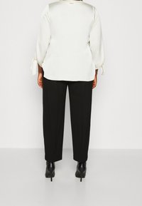 Weiße, strukturierte Bluse mit langen Ärmeln und seitlichen Bindebändern, kombiniert mit schwarzen, taillierten Hosen und schwarzen High Heels. Rückansicht des Outfits.