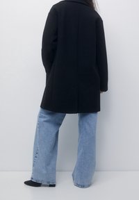 Abrigo de lana negro con cuello, corte recto y mangas largas, combinado con jeans de pierna ancha color azul claro y botines negros.
