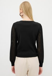 Schwarzer Pullover mit geripptem Körper und transparenten, gestreiften langen Ärmeln. Mit einem runden Halsauschnitt und eng anliegendem Bündchen. Kombiniert mit beigen Hosen.