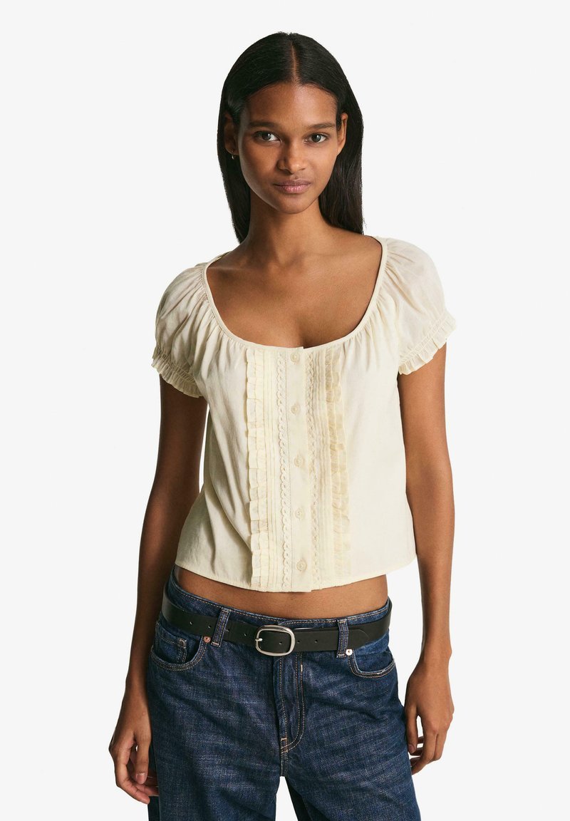 Hellbeige cropped Bluse mit kurzen, ballonförmigen Ärmeln, die ein vertikales, gerafftes Paneel und eine Knopfleiste an der Vorderseite aufweist. Getragen mit dunklen Jeans.