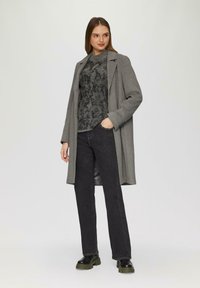 Manteau gris long sur un pull à motif sombre, associé à un jean noir et des bottes noires chunky. Tissu lisse avec une coupe ajustée.