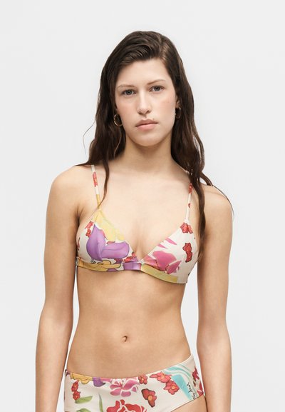 Jeune femme aux longs cheveux bruns portant un bikini fleuri coloré, debout devant un fond clair uni.