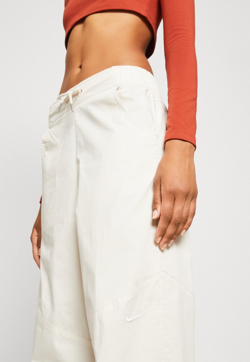 Beige weite Damen-Hosen von Nike mit elastischem Bund, Seitentaschen, dezentem Stich und kleinem weißen Logo am linken Bein.
