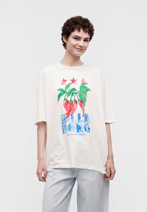 Jeune personne aux cheveux courts et bouclés porte un T-shirt blanc oversize avec des piments rouges, des feuilles vertes, trois étoiles rouges et le texte bleu "Billabong".