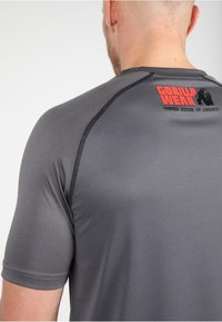 Camisa gris de manga corta hecha de una tela suave, con un logotipo rojo y negro en la parte trasera y costuras contrastantes en negro a lo largo de las costuras.