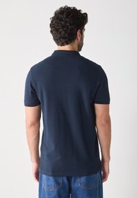 Camiseta polo azul marino con mangas cortas, hecha de una suave tela de algodón. Presenta un collar clásico y un corte recto, se muestra la vista trasera.