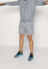 Sudadera deportiva gris y pantalones cortos hechos de una tela suave, con cierre frontal de cremallera y bolsillos. El hombre lleva unas zapatillas azules con detalles negros.