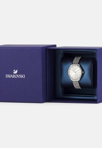 Sølvtoneur med en rund urskive, krystalaccenter på en mesh-rem, præsenteret i en marineblå boks med en tekstureret overflade.