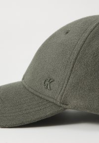 Casquette de baseball grise avec une visière incurvée, arborant un logo "CK" ton sur ton à l'avant et des détails de couture visibles.