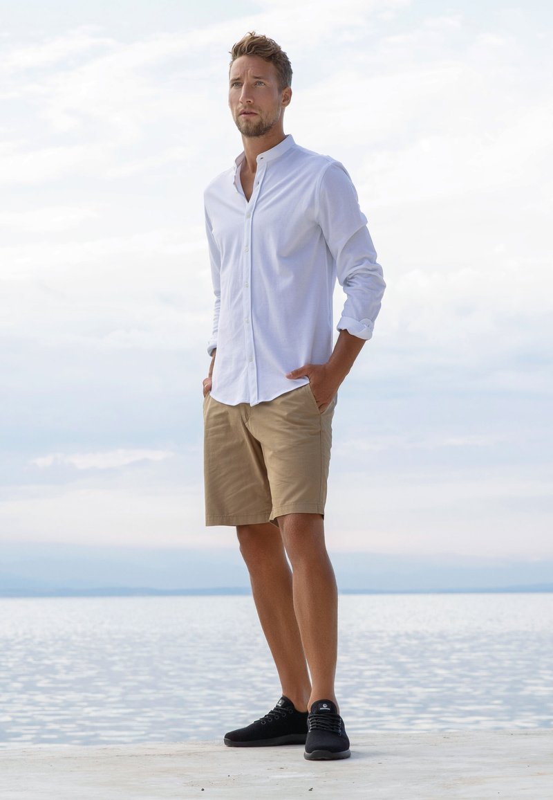 White button-up shirt, khaki shorts, black sneakers. Model standing with hands in pockets against a water backdrop with cloudy sky.