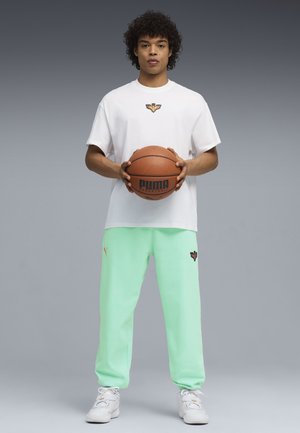 Młody mężczyzna stoi trzymając piłkę do koszykówki, ubrany w biały T-shirt i miętowo-zielone spodnie dresowe na jednolitym szarym tle.