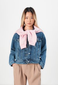 Denim jacket, blue, with button front and two chest pockets, featuring a light pink sweatshirt tied around the neck. Trousers are beige.
