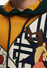 Kapuzensweatshirt in senfgelb. Mit einem großen grafischen Design von Zeichentrickfiguren. Kontrastierende dunkelgrüne Kapuze und marineblaue Zugbänder.