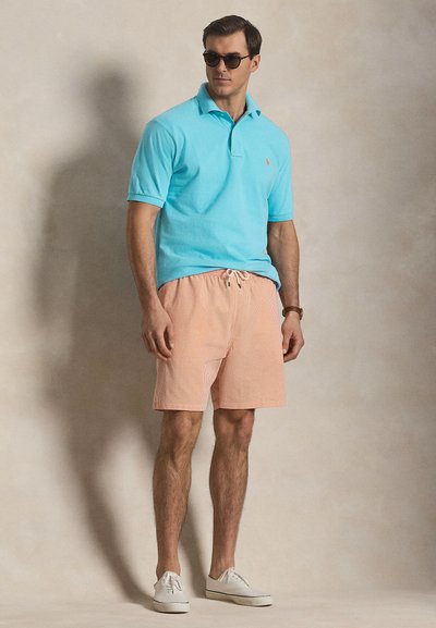 Άνδρας που φοράει τουρκουάζ πόλο, πορτοκαλί ριγωτές σορτς, λευκά αθλητικά παπούτσια, γυαλιά ηλίου και ρολόι, στέκεται μπροστά σε απλό μπεζ φόντο.