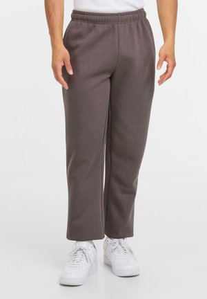 Person iført brune lige bukser og hvide sneakers, der står foran en ensfarvet lys baggrund.