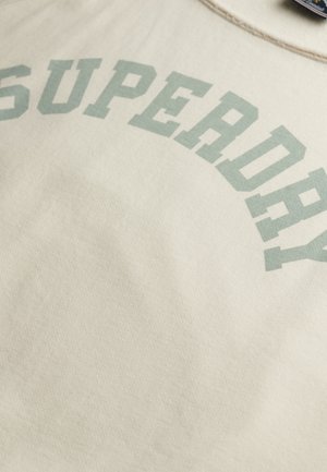 Zbliżenie beżowej tkaniny z nadrukiem „SUPERDRY” dużymi, jasnozielonymi literami w kształcie łuku.