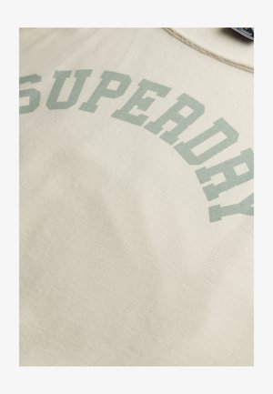 Zbliżenie beżowej tkaniny z nadrukiem „SUPERDRY” dużymi, jasnozielonymi literami w kształcie łuku.