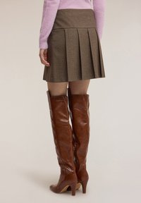 Minigonna plissettata a quadri marroni con vita alta, abbinata a stivali alti marroni in pelle con tacco che arrivano a metà coscia, caratterizzati da una texture lucida.