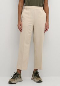 Pantaloni beige su misura con una superficie liscia, dotati di pieghe frontali e taglio dritto, abbinati a sneakers chunky beige e verdi.