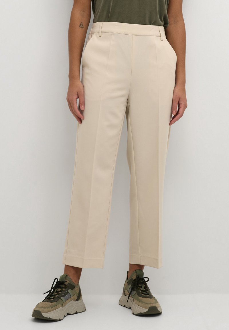 Pantaloni beige su misura con una superficie liscia, dotati di pieghe frontali e taglio dritto, abbinati a sneakers chunky beige e verdi.
