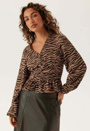 Kvinde med krøllet hår iført brun og sort zebra-mønstret bluse med lange ærmer og mørkegrøn højhøjestet nederdel, poserende mod en lys baggrund.