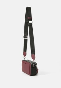 Petit sac bandoulière avec un devant bordeaux, des côtés noirs, une bandoulière noire réglable, une quincaillerie en argent et une fermeture éclair double.
