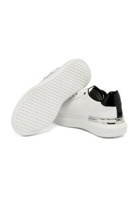 Sneaker bianco con un accento nero sul tallone, caratterizzato da una striscia argentata lucida. La suola presenta un grip testurizzato e decorato.