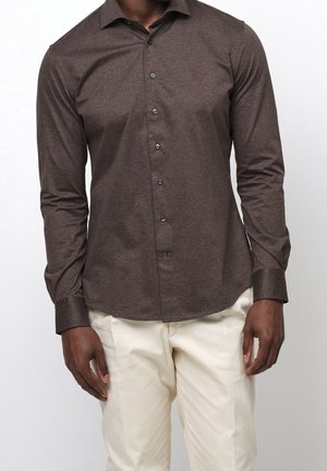 Mann trägt ein eng anliegendes dunkelbraunes Langarm-Hemd mit Knöpfen und cremefarbene Hose, steht vor einem einfarbigen Hintergrund.