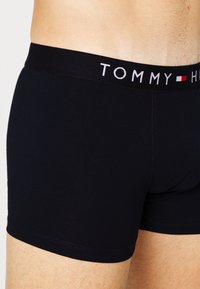 Juodi medvilniniai boxeriai su prigludusia forma, turintys kontrastingą juosmens juostą su "TOMMY" prekės ženklu ir raudonais bei baltais akcentais.