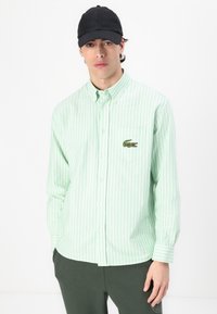 Light green striped button-up shirt with a logo patch on the chest, paired with dark green pants; fabric appears smooth and breathable.